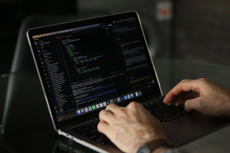 A person's hands typing on a sleek, modern laptop with a black screen displaying lines of colorful code, placed on a dark reflective surface. The laptop’s keyboard is black with white lettering, and